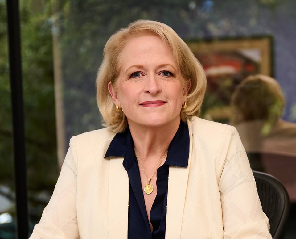 Ellen Wood, a professional woman with blonde hair, wearing a cream blazer and navy blouse, seated in an office with a window and artwork behind her.
